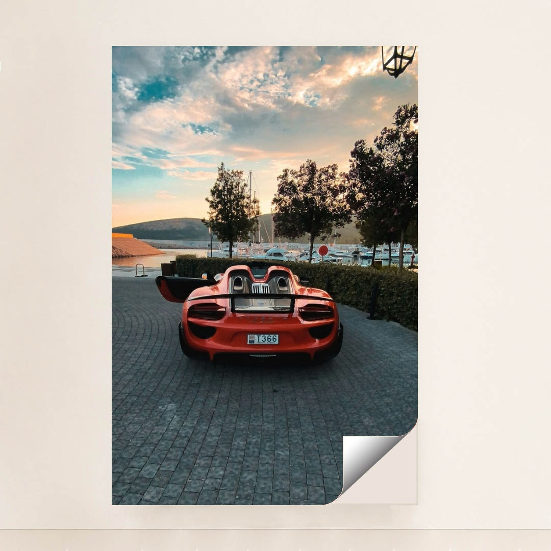 This photo shows a red Porsche 918 Spyder with open doors parked on a cobbled marina at sunset, unframed style.