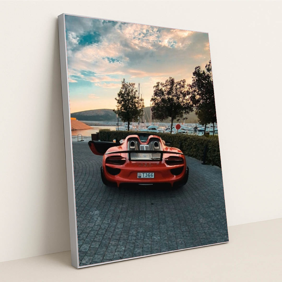 This photo shows a red Porsche 918 Spyder with open doors parked on a cobbled marina at sunset, in a silver frame.