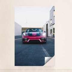 This photo shows a pink and black Porsche sports car viewed from the front, parked on an urban street, unframed.