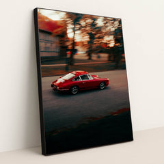 This photo shows a red vintage Porsche sports car driving through a city street at sunset, in a black frame.