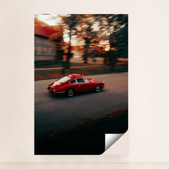 This photo shows a red vintage Porsche sports car driving through a city street at sunset, minimalist unframed style.