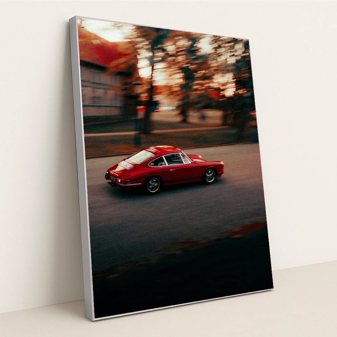 This photo shows a red vintage Porsche sports car driving through a city street at sunset, in a silver frame.