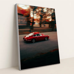 This photo shows a red vintage Porsche sports car driving through a city street at sunset, in a silver frame.