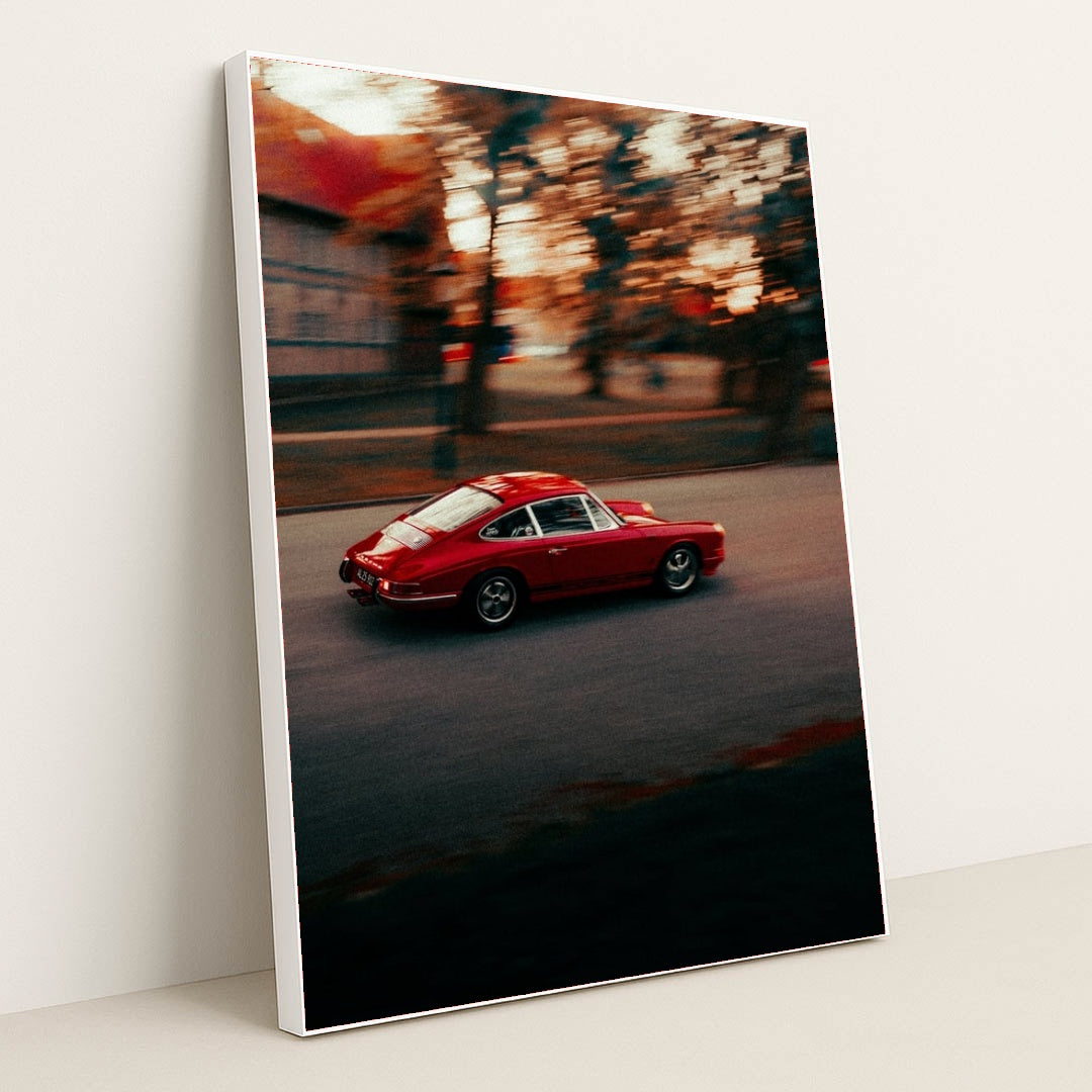 This photo shows a red vintage Porsche sports car driving through a city street at sunset, in a white frame.
