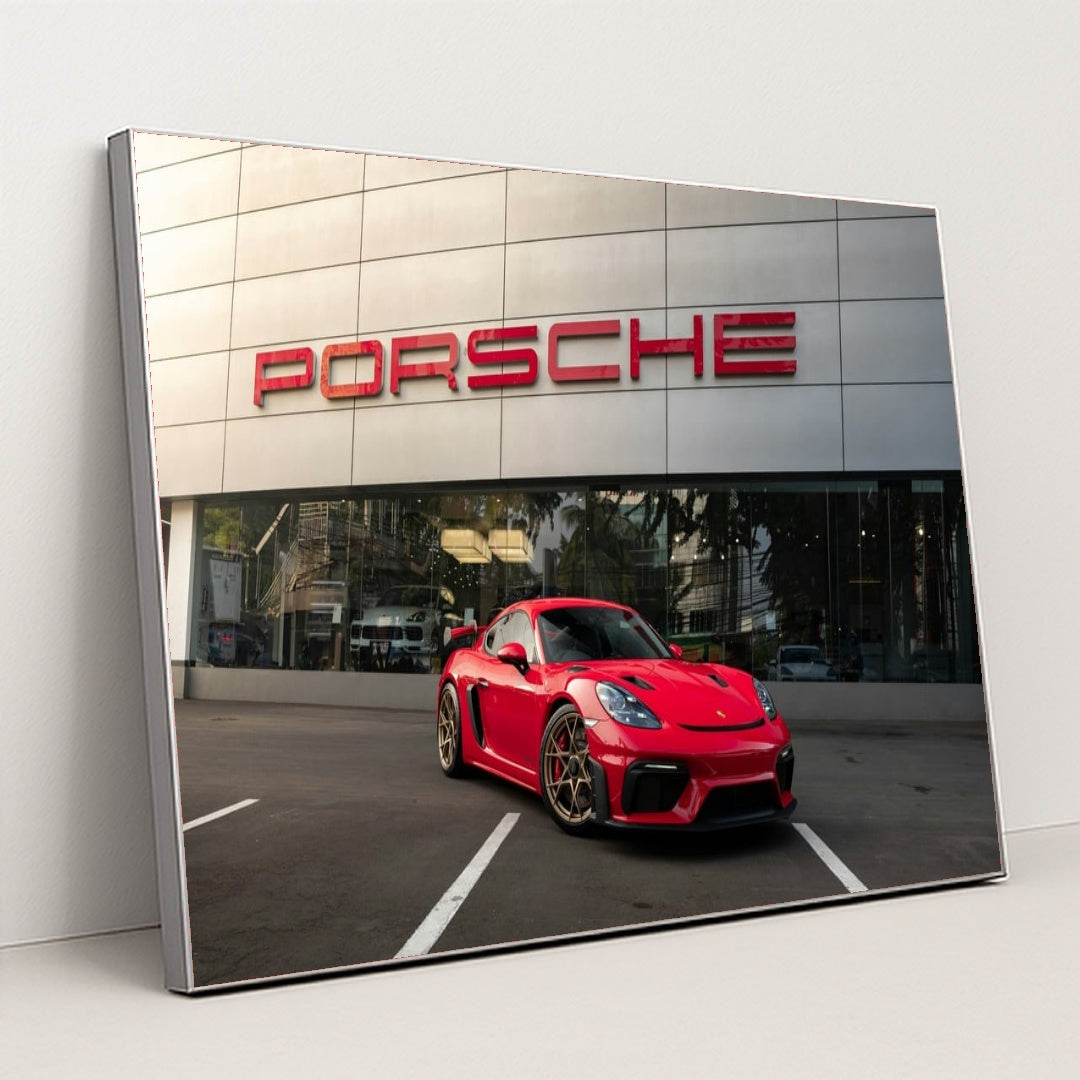 This photo shows a red Porsche sports car parked in front of a modern dealership building with a large Porsche sign, in a silver frame.
