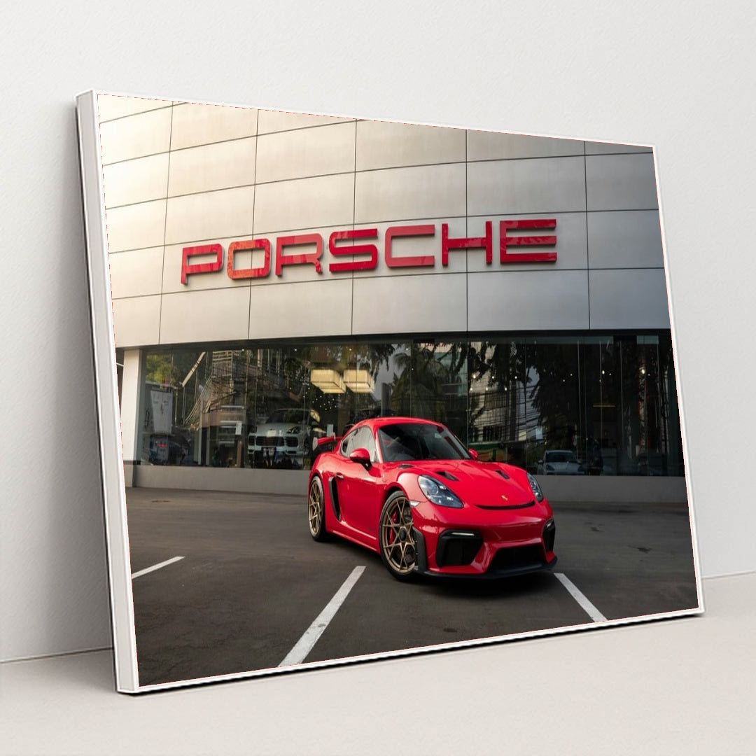 This photo shows a red Porsche sports car parked in front of a modern dealership building with a large Porsche sign, in a white frame.