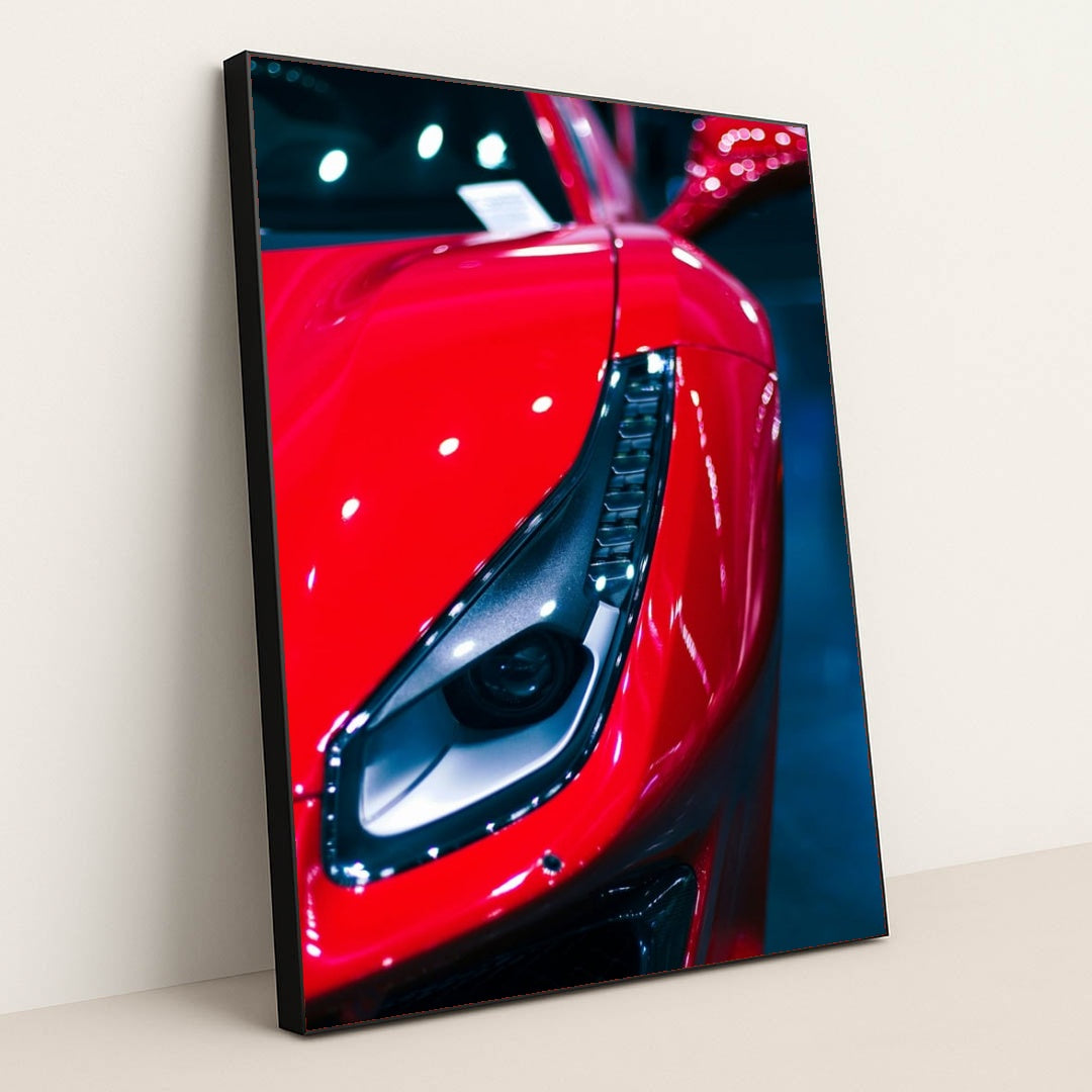This photo shows a close-up of the left headlight and front fender of a red Ferrari under bright lights, in a black frame.