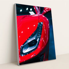 This photo shows a close-up of the left headlight and front fender of a red Ferrari under bright lights, in a silver frame.