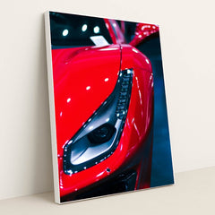 This photo shows a close-up of the left headlight and front fender of a red Ferrari under bright lights, in a white frame.