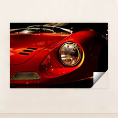 This photo shows a close-up of the front left headlight and fender of a classic red Ferrari Dino, unframed style.