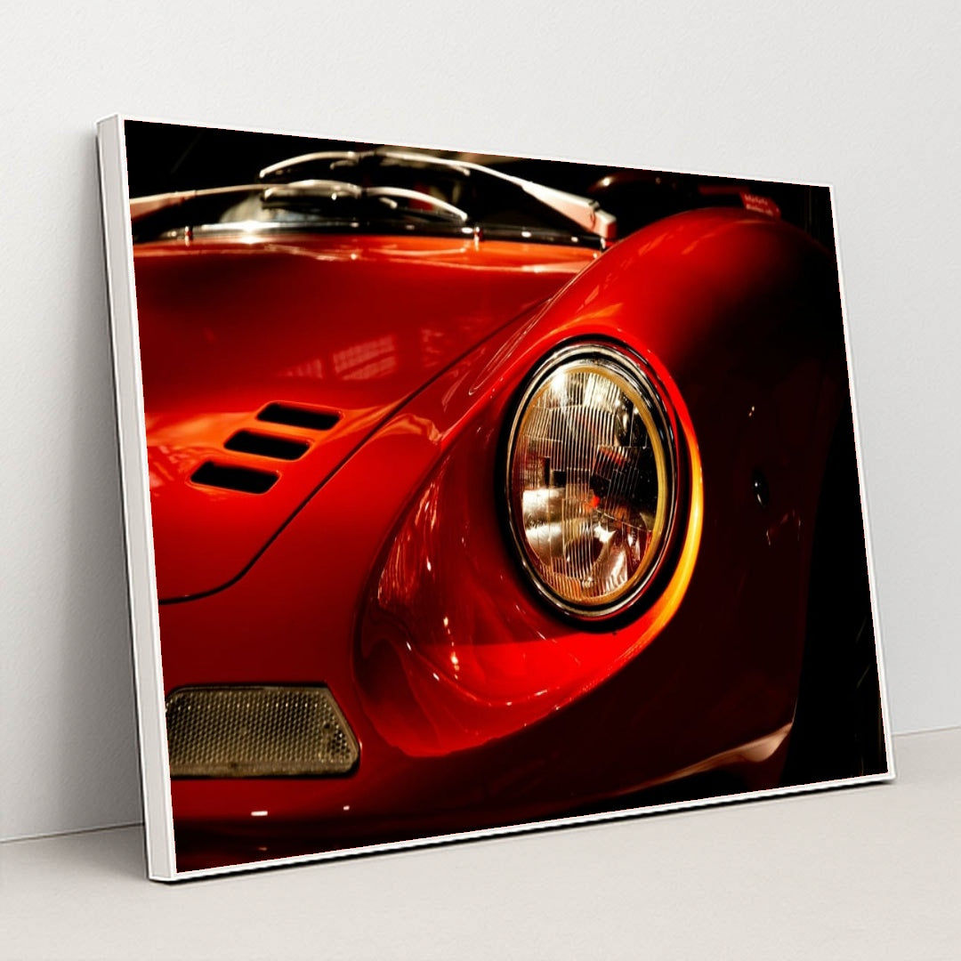 This photo shows a close-up of the front left headlight and fender of a classic red Ferrari Dino, in a white frame.