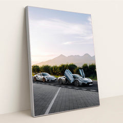This photo shows a white Porsche sports car and a silver supercar with open butterfly doors parked by the mountains at sunrise, in a silver frame.