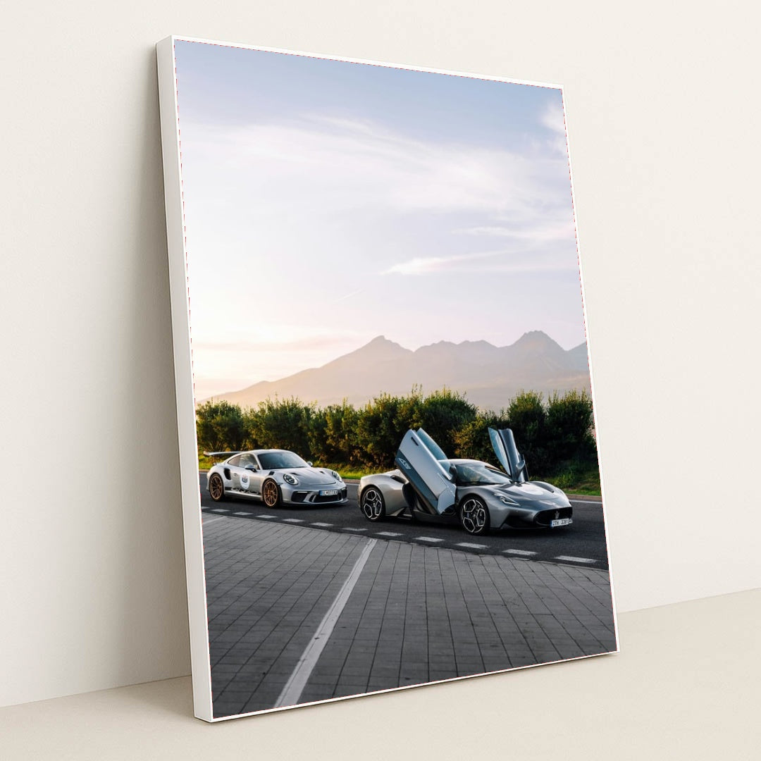 This photo shows a white Porsche sports car and a silver supercar with open butterfly doors parked by the mountains at sunrise, in a white frame.