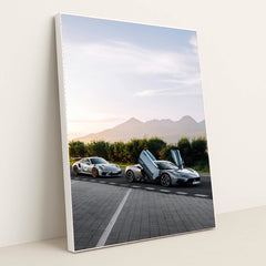 This photo shows a white Porsche sports car and a silver supercar with open butterfly doors parked by the mountains at sunrise, in a white frame.