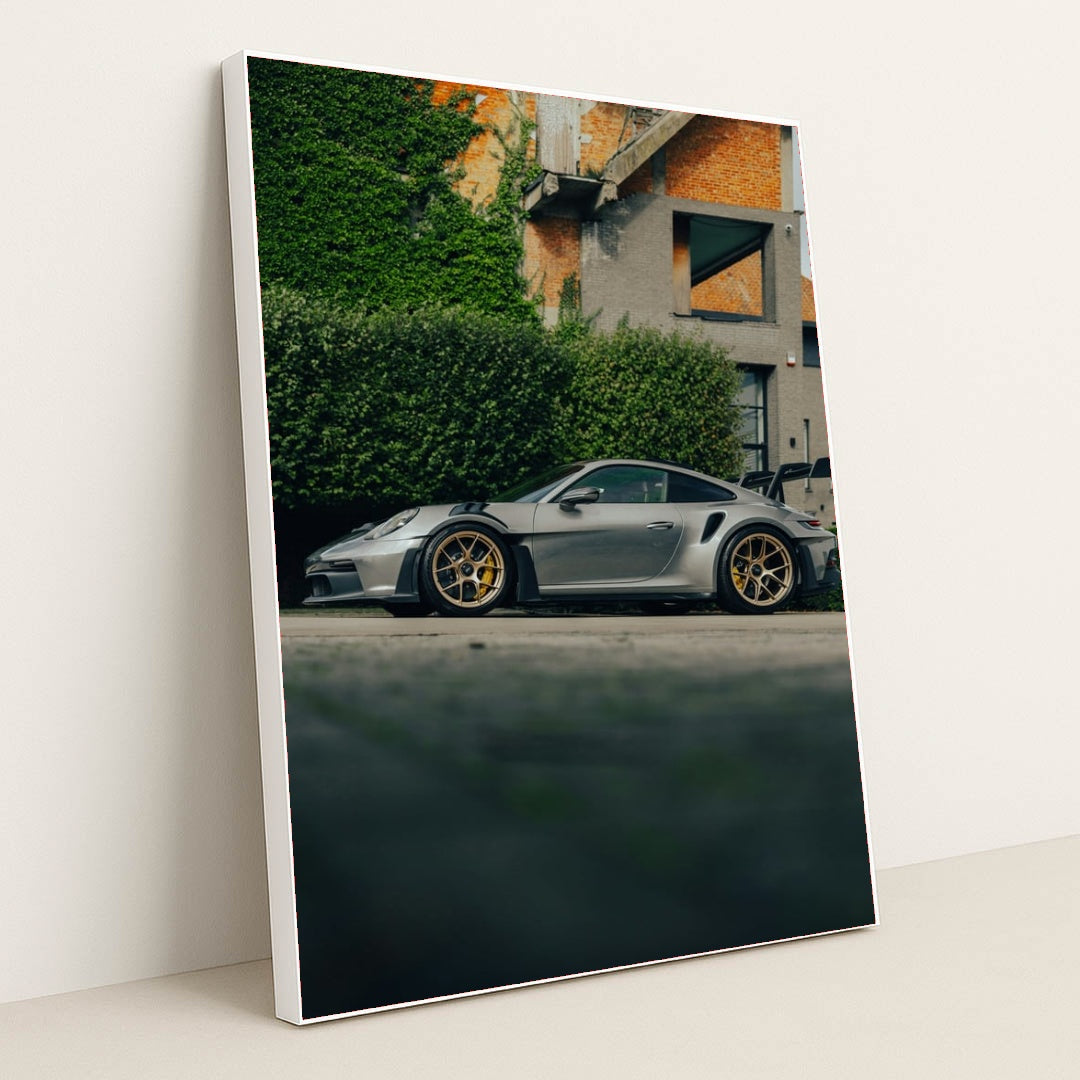 This photo shows a silver Porsche 911 GT3 RS with gold wheels parked beside a hedge and brick building, in a white frame.