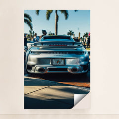 This photo shows the rear of a silver Porsche 911 Turbo S parked outdoors with palm trees and people in the background, unframed.