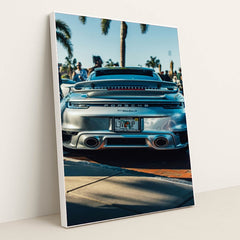 This photo shows the rear of a silver Porsche 911 Turbo S parked outdoors with palm trees and people in the background, in a white frame.