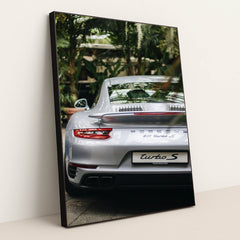 This photo shows the rear view of a silver Porsche 911 Turbo S parked among lush green plants, in a black frame.