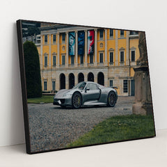This photo shows a silver Porsche 918 Spyder parked in front of an elegant yellow European mansion with banners, in a black frame.