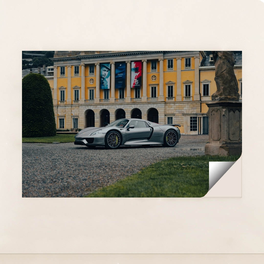 This photo shows a silver Porsche 918 Spyder parked in front of an elegant yellow European mansion with banners, unframed style.