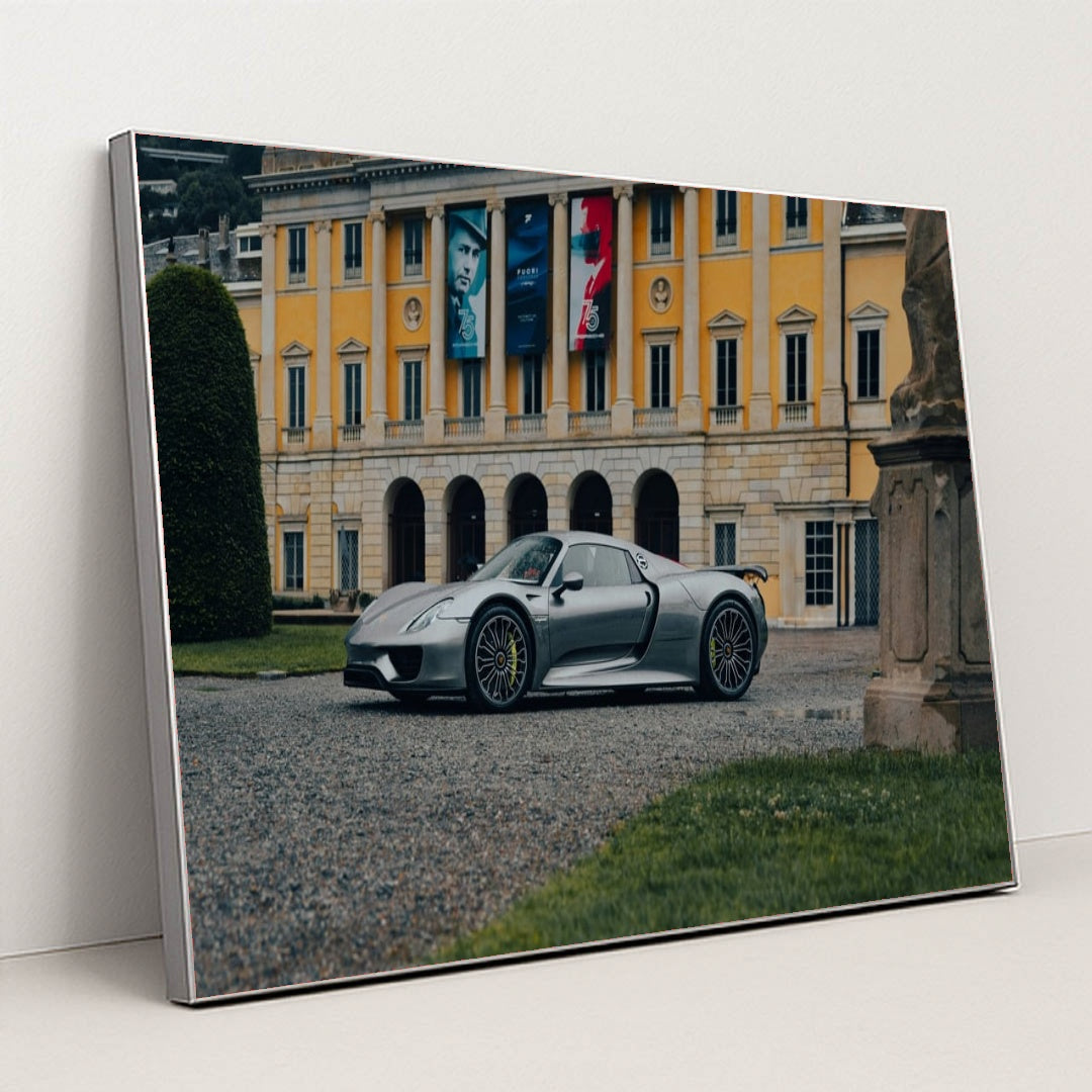 This photo shows a silver Porsche 918 Spyder parked in front of an elegant yellow European mansion with banners, in a silver frame.