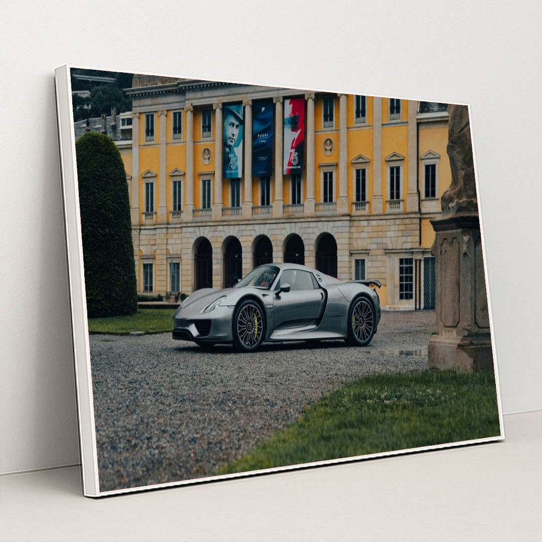 This photo shows a silver Porsche 918 Spyder parked in front of an elegant yellow European mansion with banners, in a white frame.