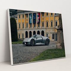 This photo shows a silver Porsche 918 Spyder parked in front of an elegant yellow European mansion with banners, in a white frame.