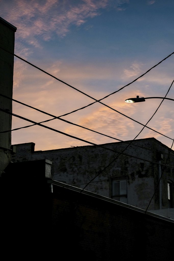 Connor McManus - Silhouetted urban buildings with intersecting wires against a colorful dusk sky with clouds.