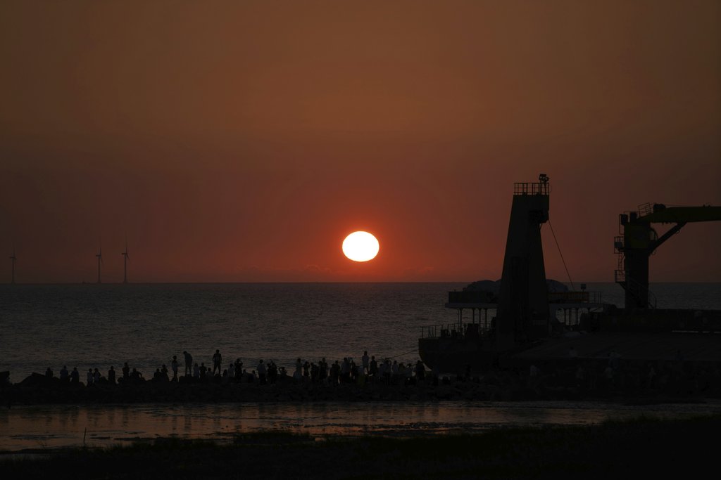 Roy - A vibrant sunset casts silhouettes over a harbor with wind turbines in view.