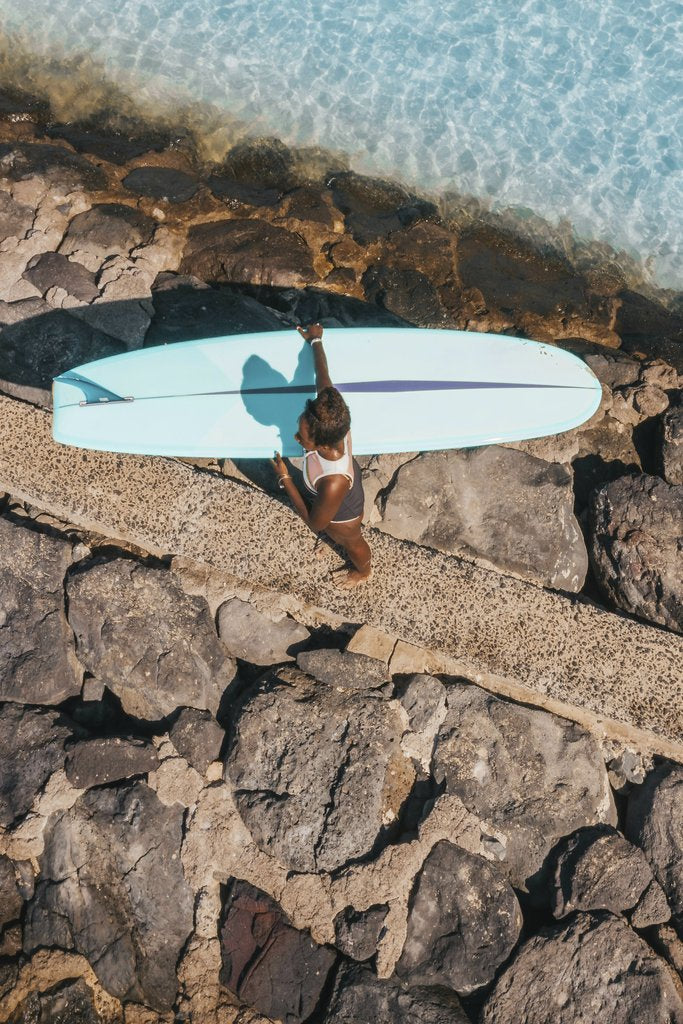 Jess Loiterton - A surfer walking along a rocky path with a surfboard near water in a sunny setting.