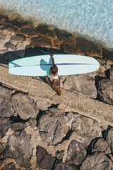 Jess Loiterton - A surfer walking along a rocky path with a surfboard near water in a sunny setting.