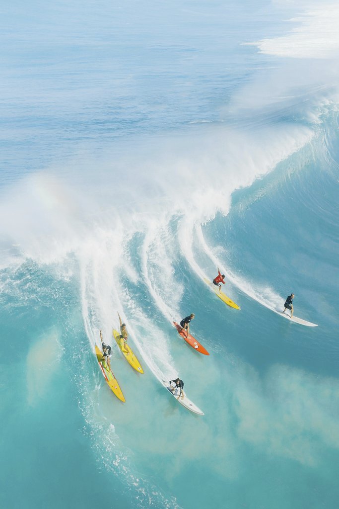 Jess Loiterton - Dynamic aerial photo capturing surfers conquering massive waves in vibrant blue Hawaiian waters.