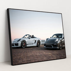 This photo shows a white convertible Porsche and a black coupe Porsche parked together at sunrise, in a black frame.