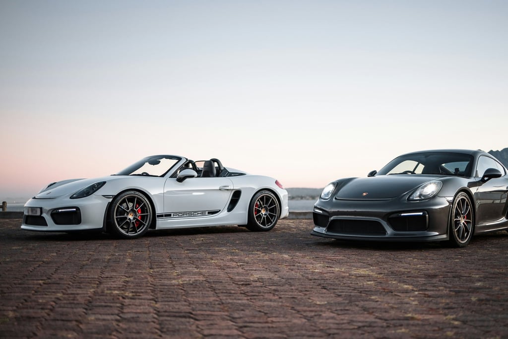 Porsche Duo at Dusk: white Boxster Spyder and graphite Cayman GT4 on rustic pavement under dusk sky, showcasing car wall art.