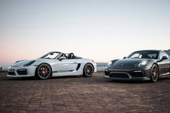 Porsche Duo at Dusk: white Boxster Spyder and graphite Cayman GT4 on rustic pavement under dusk sky, showcasing car wall art.