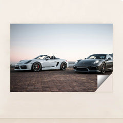 This photo shows a white convertible Porsche and a black coupe Porsche parked together at sunrise, unframed.