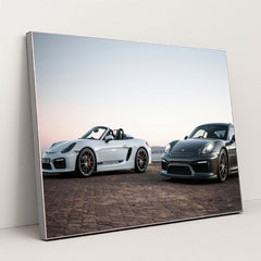 This photo shows a white convertible Porsche and a black coupe Porsche parked together at sunrise, in a silver frame.