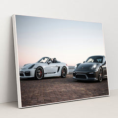 This photo shows a white convertible Porsche and a black coupe Porsche parked together at sunrise, in a white frame.