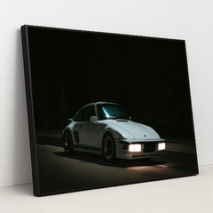 This photo shows a white Porsche 911 with pop-up headlights and wide body kit driving at night, in a black frame.