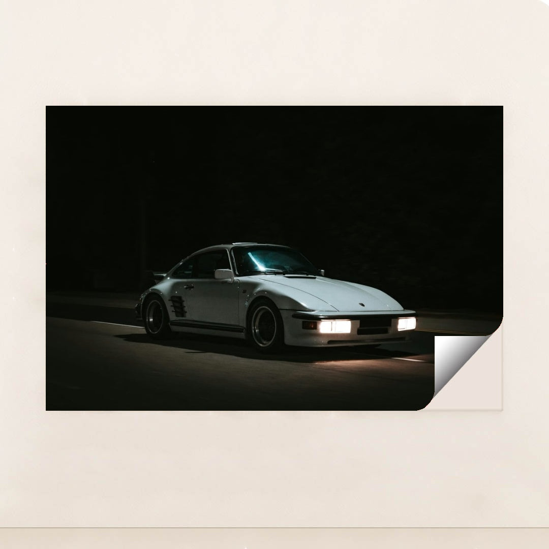 This photo shows a white Porsche 911 with pop-up headlights and wide body kit driving at night, minimalist unframed style.