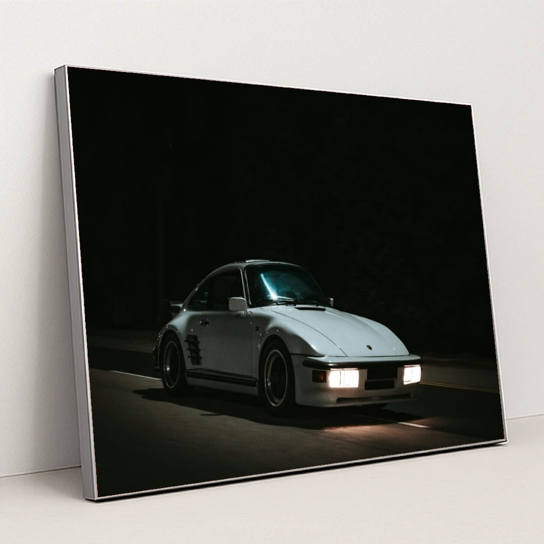 This photo shows a white Porsche 911 with pop-up headlights and wide body kit driving at night, in a silver frame.