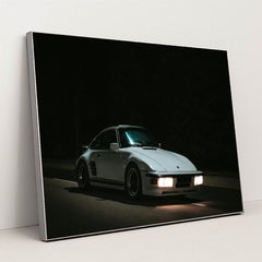 This photo shows a white Porsche 911 with pop-up headlights and wide body kit driving at night, in a silver frame.
