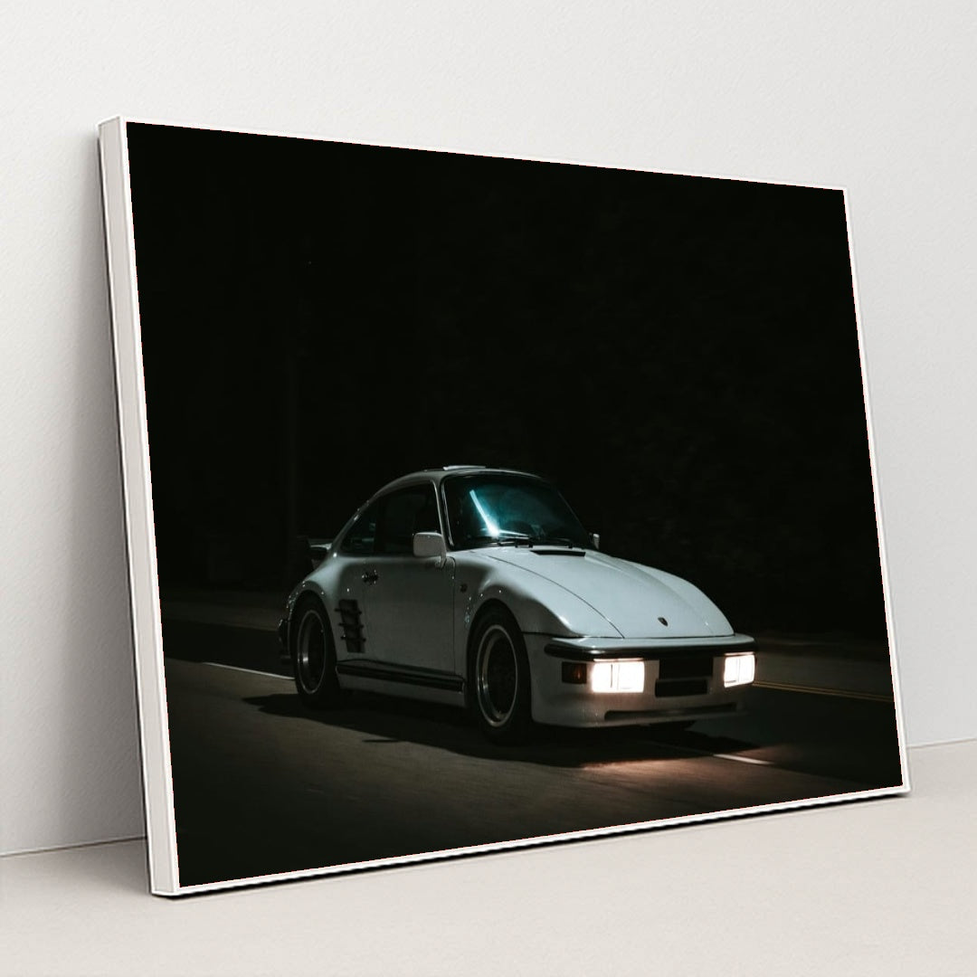 This photo shows a white Porsche 911 with pop-up headlights and wide body kit driving at night, in a white frame.