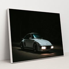 This photo shows a white Porsche 911 with pop-up headlights and wide body kit driving at night, in a white frame.