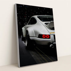 This photo shows the rear of a white Porsche sports car with a large wing, illuminated taillights, and starry reflections, in a silver frame.