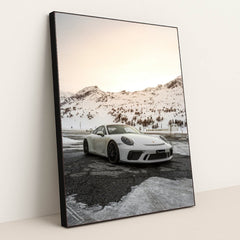 This photo shows a white Porsche sports car parked on a snow-covered mountain road at sunset, in a black frame.