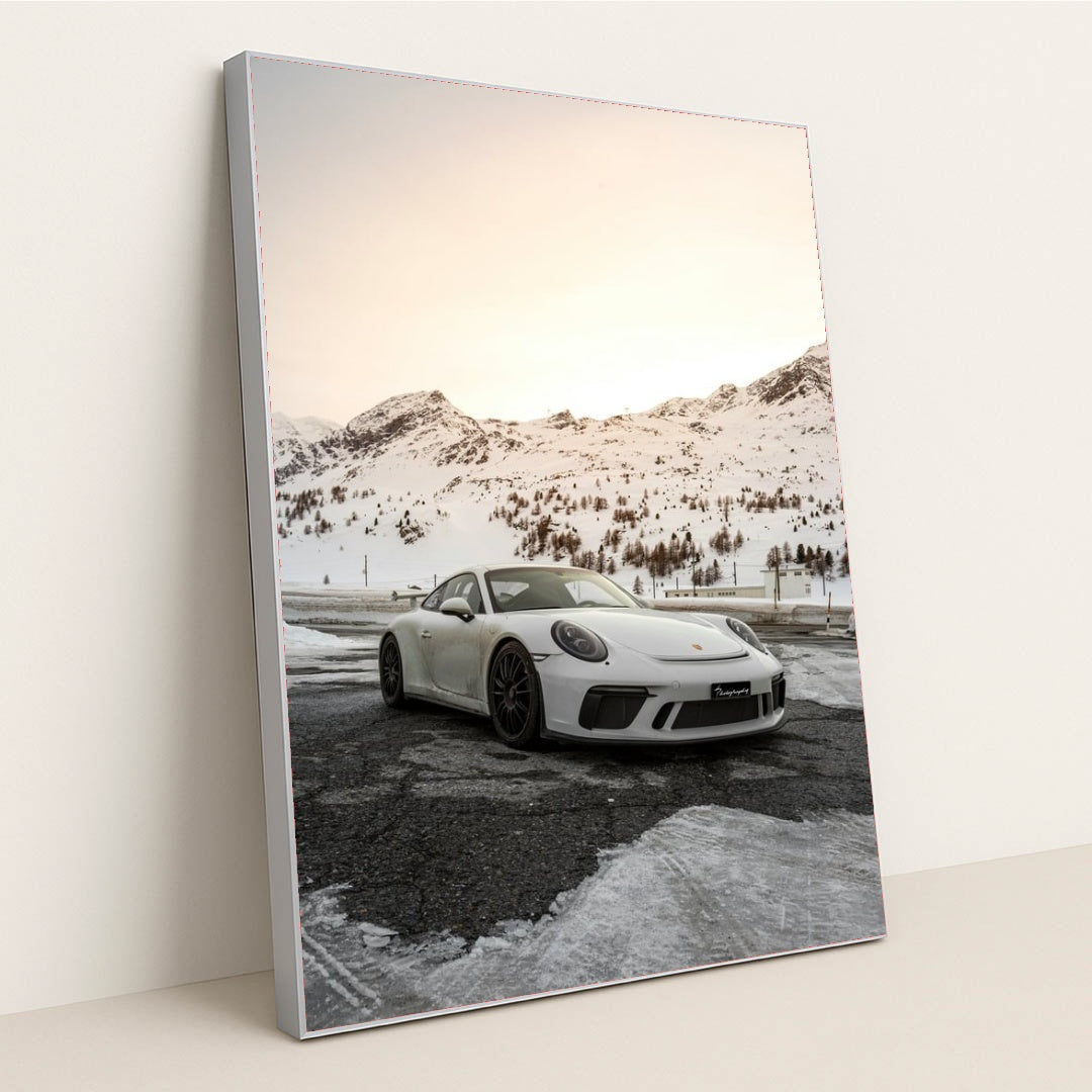 This photo shows a white Porsche sports car parked on a snow-covered mountain road at sunset, in a silver frame.