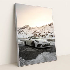 This photo shows a white Porsche sports car parked on a snow-covered mountain road at sunset, in a silver frame.