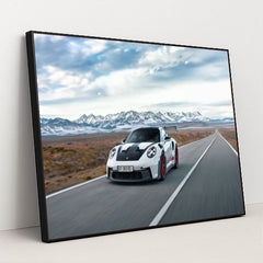 This photo shows a white Porsche 911 GT3 RS with red wheels driving quickly on an open desert highway with mountains in the background, in a black frame.