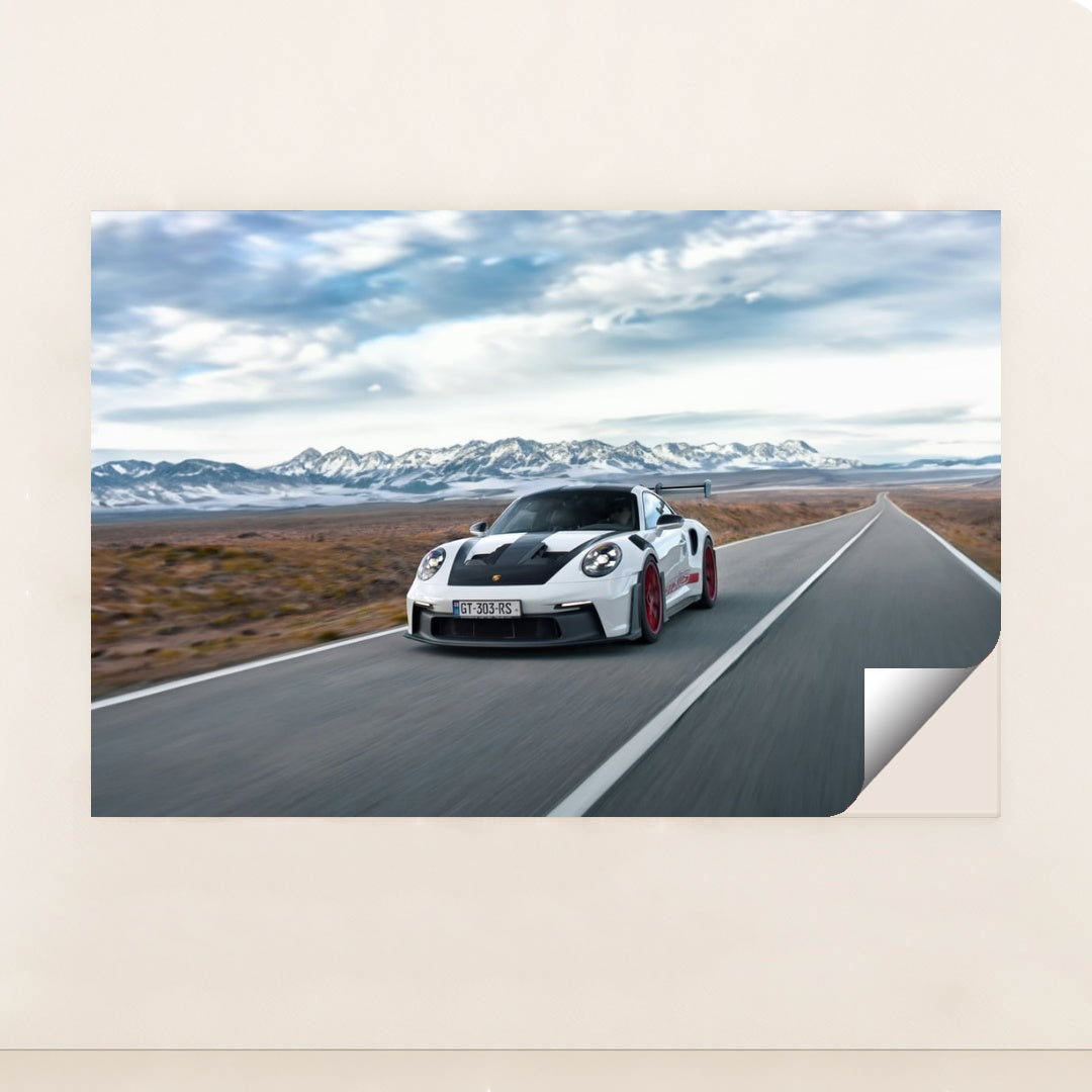 This photo shows a white Porsche 911 GT3 RS with red wheels driving quickly on an open desert highway with mountains in the background, unframed style.